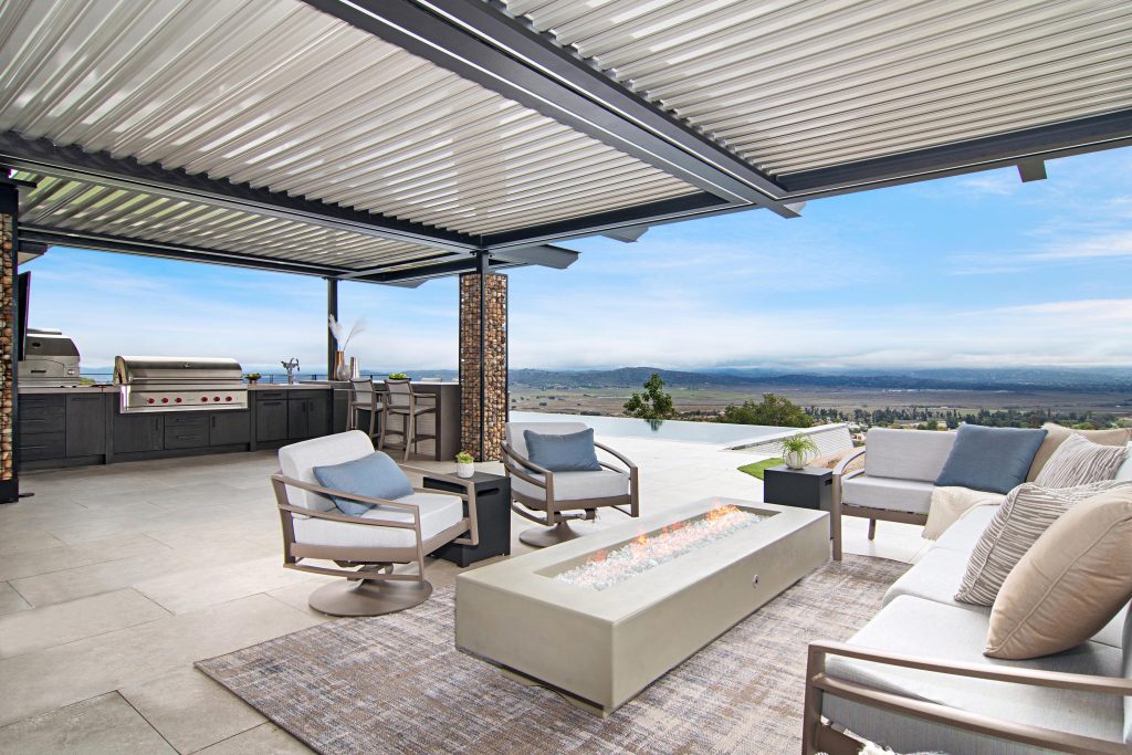 A white pergola with a seating area and a kitchen on the side overlooking the plains and the louvered pergola is closed for rain protection